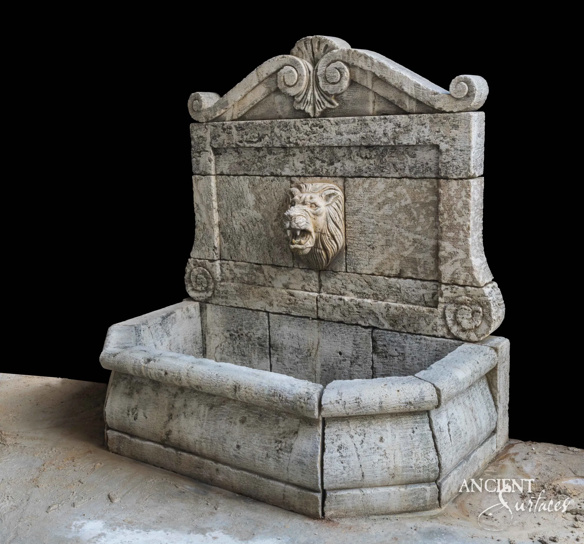 Reclaimed antique limestone wall fountain from Ancient Surfaces with an arched scrolled crest, a carved lion mask spout, and a canted rectangular basin, photographed against black