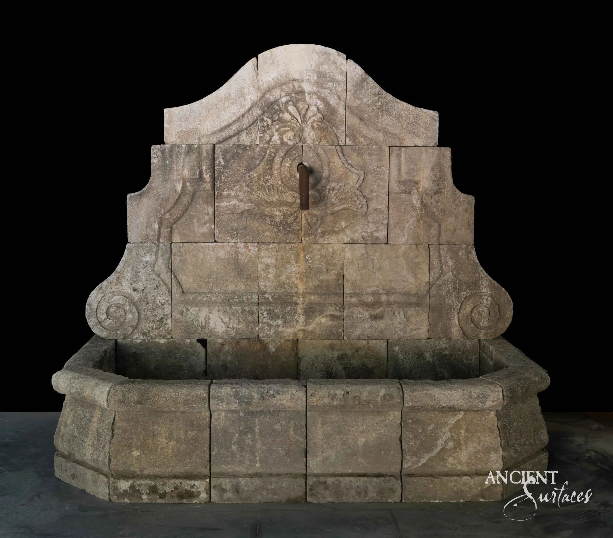Grand reclaimed antique limestone wall fountain from Ancient Surfaces with an arched carved crest, a shield cartouche fitted with an iron spout, and a wide canted basin, photographed against black