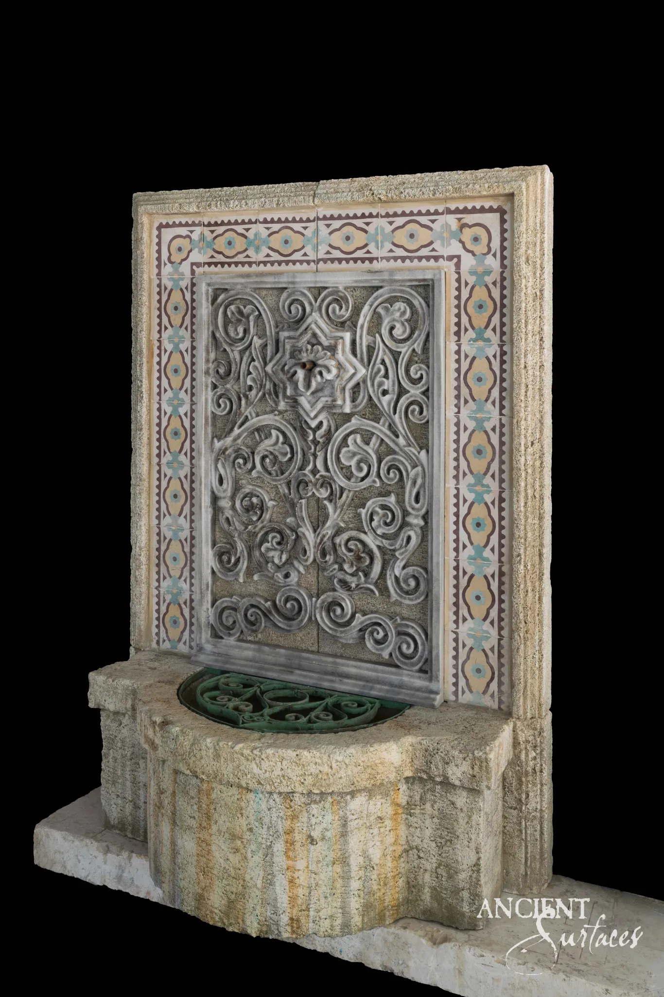 Reclaimed antique limestone wall fountain from Ancient Surfaces framed in Moorish style mosaic tile with a scrolled metal grille panel and a shaped basin fitted with a bronze floor grate