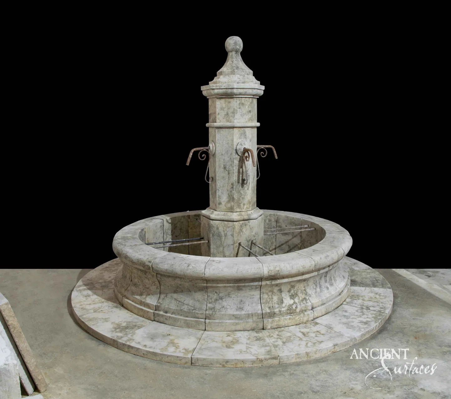 Reclaimed antique limestone pedestal fountain from Ancient Surfaces with a fluted column, a ball finial, twin scrolled iron spouts, and a round basin on a stepped stone platform, photographed against black