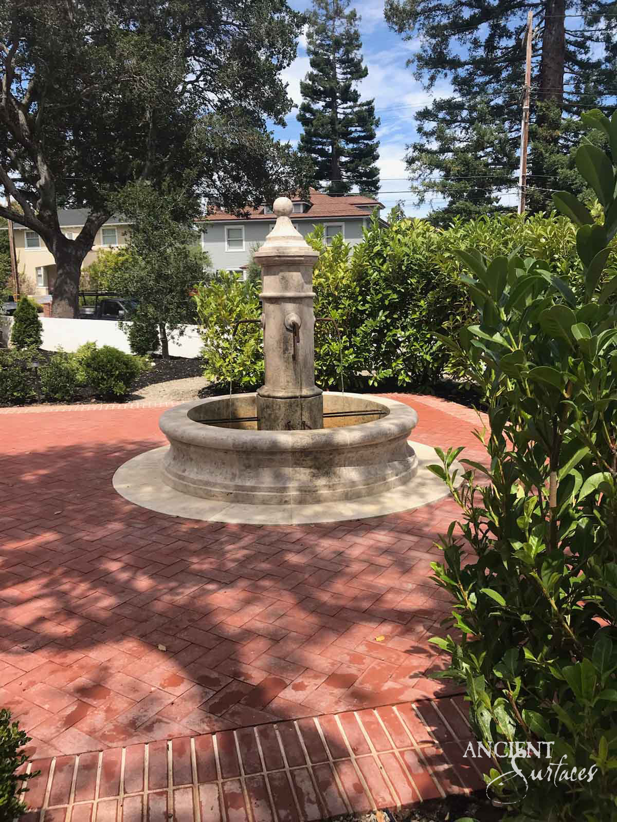 Reclaimed antique limestone pedestal fountain from Ancient Surfaces with a ball finial and round basin, installed at the centre of a herringbone brick garden patio surrounded by mature trees and hedges