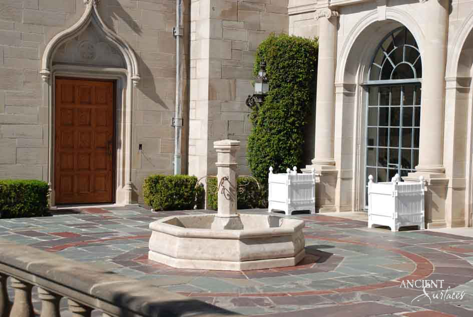 Reclaimed antique limestone octagonal basin fountain from Ancient Surfaces on a stepped plinth in a grand stone entrance courtyard, flanked by an arched doorway and clipped topiary in white planters