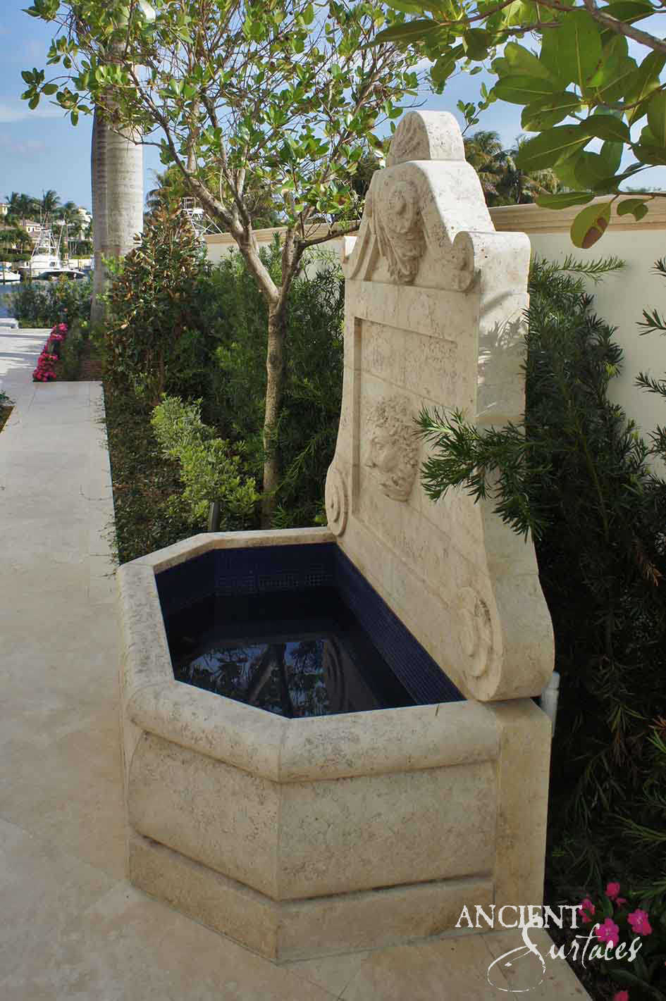 Tall reclaimed antique limestone wall fountain from Ancient Surfaces with a carved medallion backplate and a hexagonal basin lined in dark tile, mounted on a cream stucco wall among palm trees