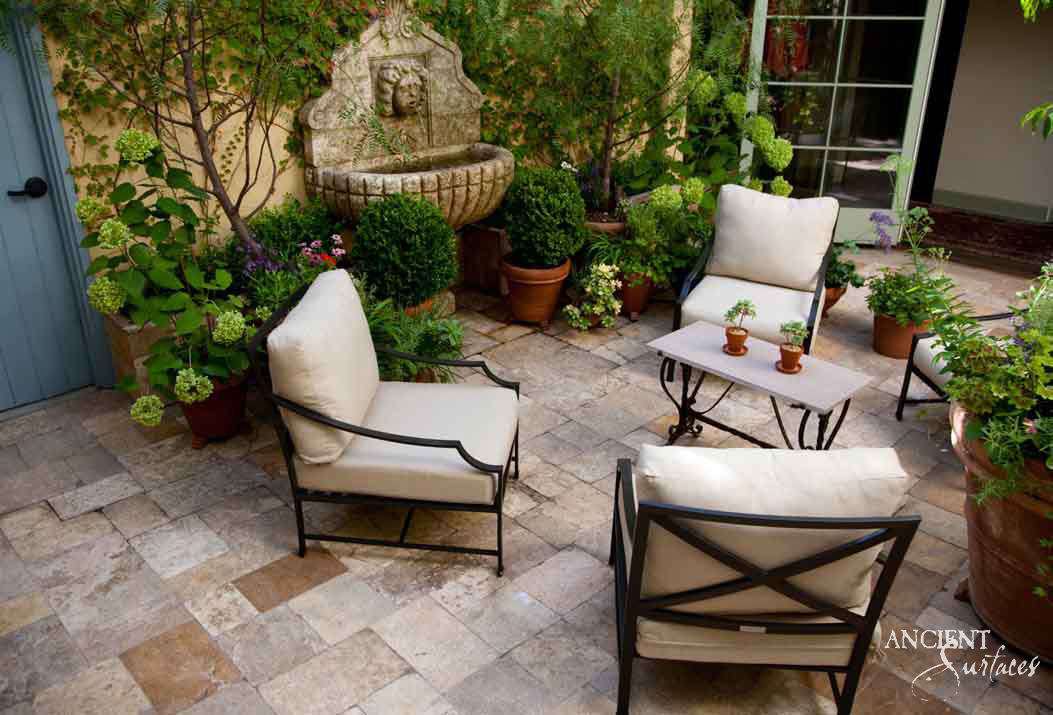 Reclaimed antique limestone lion mask wall fountain from Ancient Surfaces with a shell shaped basin, mounted on an ochre plaster wall on a sunlit travertine terrace furnished with outdoor lounge chairs