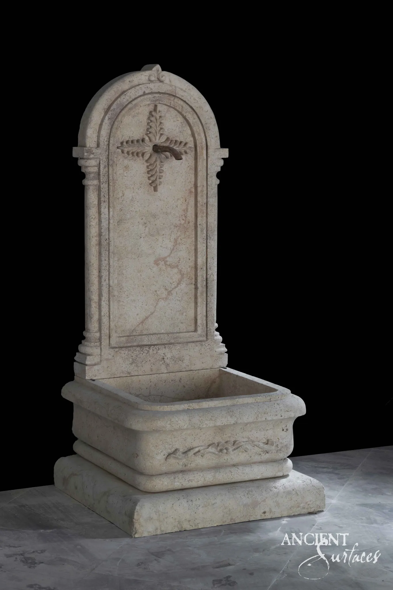 Reclaimed antique limestone Gothic wall fountain from Ancient Surfaces with a pointed arched crest carved with a quatrefoil cross above a rectangular basin, photographed against black