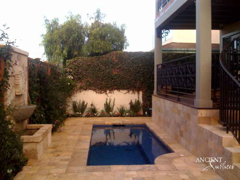 Reclaimed antique limestone wall fountain from Ancient Surfaces mounted on a garden wall and spilling into a stone trough at the edge of a terraced backyard swimming pool